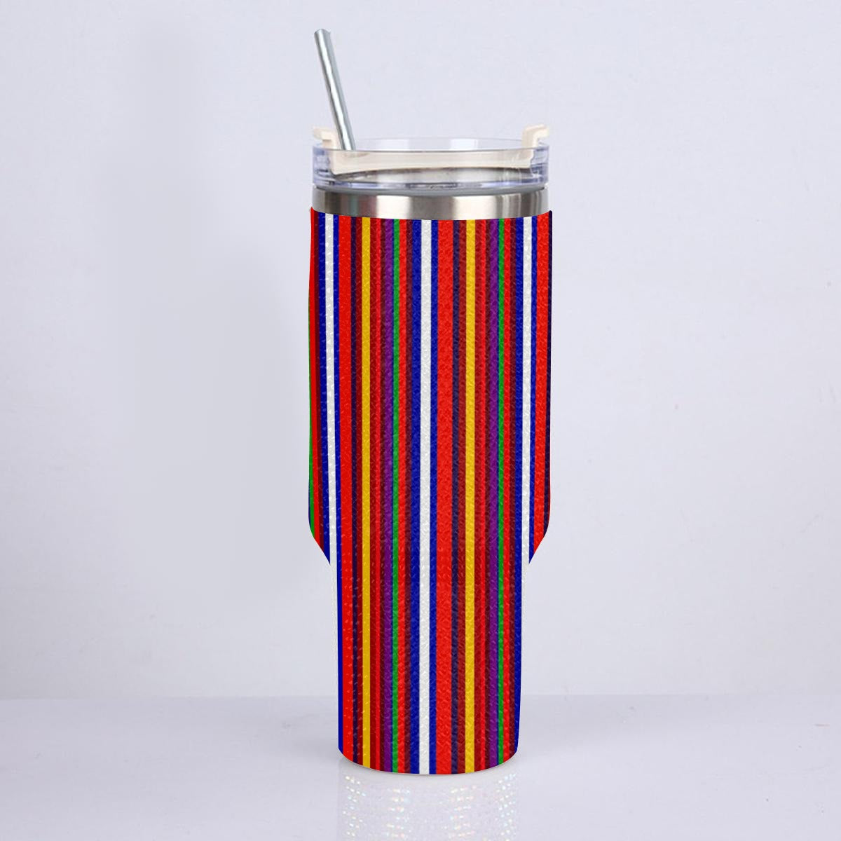 A tall insulated tumbler with a clear lid and metal straw, wrapped in a colorful vertically striped fabric sleeve featuring red, orange, yellow, green, blue, and white stripes, set against a plain light gray background.
