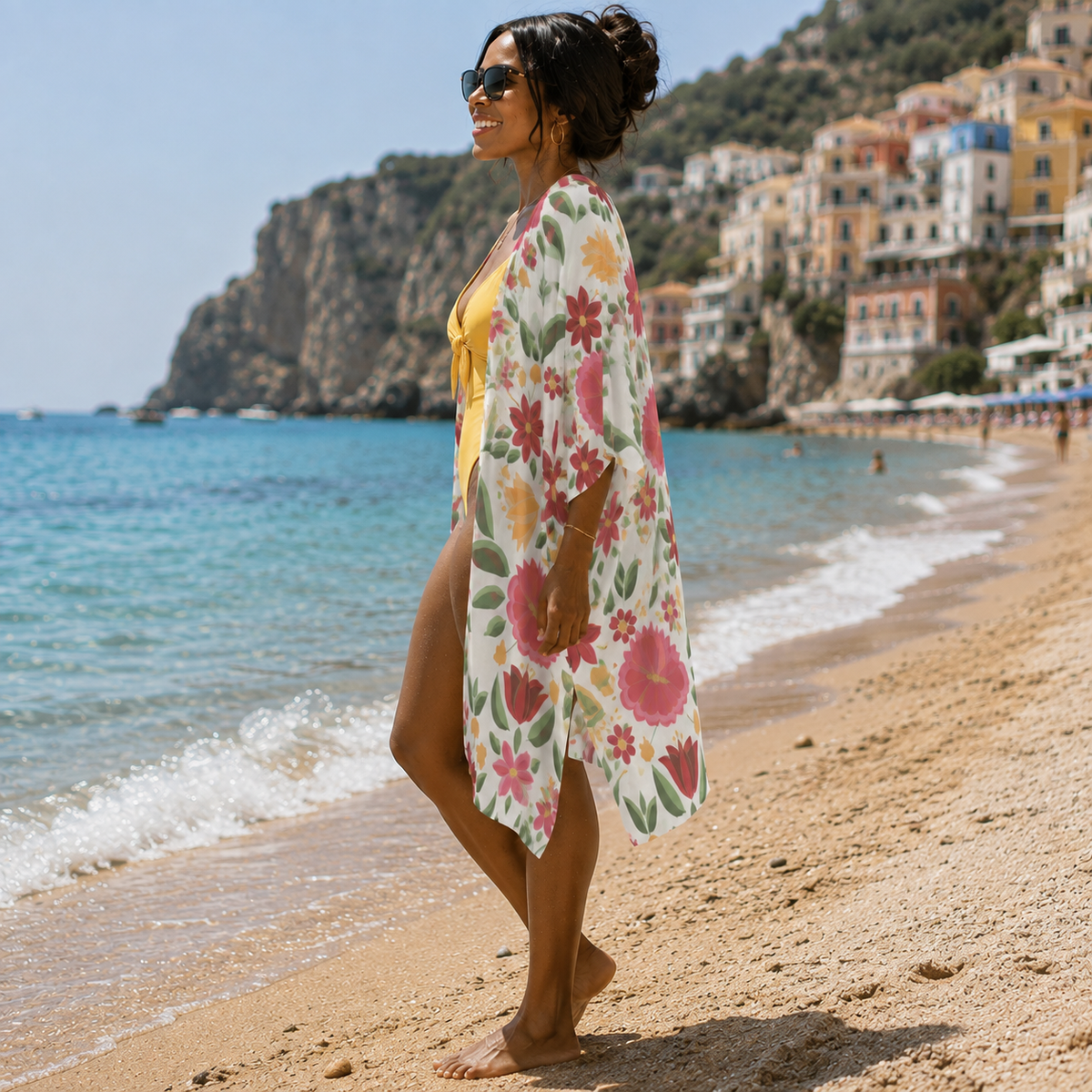 A woman stands on a sandy beach wearing a white floral kimono cover-up over a yellow bikini, with turquoise water and a hillside town in the background.