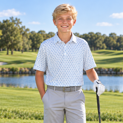 A young boy stands on a golf course holding a driver, wearing a white patterned short-sleeve polo shirt, light gray shorts, white golf shoes, and a white glove. A golf bag with clubs is visible in the background near a lake.