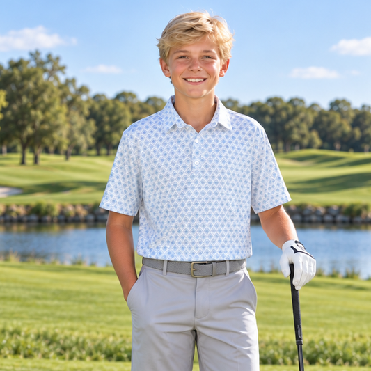 A young boy stands on a golf course holding a driver, wearing a white patterned short-sleeve polo shirt, light gray shorts, white golf shoes, and a white glove. A golf bag with clubs is visible in the background near a lake.