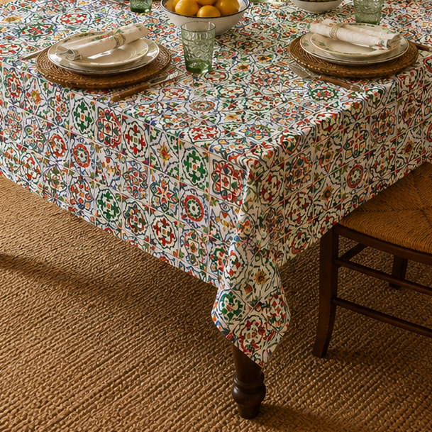 A rectangular dining table covered with a multicolor floral tablecloth, set with plates, glasses, and a vase of flowers in a warmly lit dining room with wooden chairs, a chandelier, and decorative wall art.