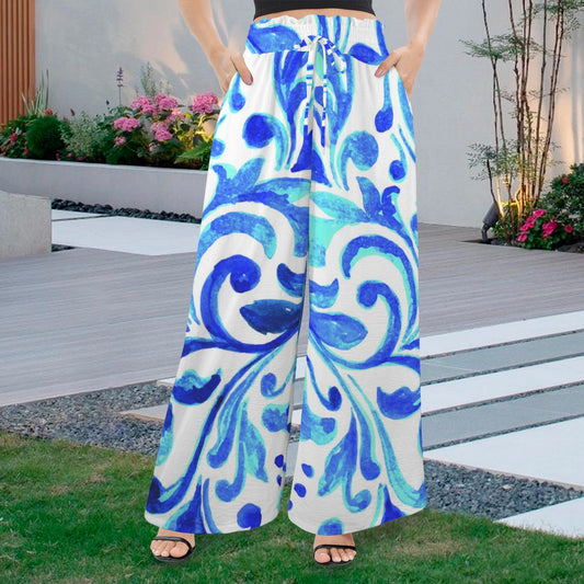 Full-body shot of a woman wearing Ondas Wide Leg Pants featuring a vibrant blue and white Mediterranean scroll print, paired with a white sleeveless top and straw purse, standing on a path at a tropical resort.
