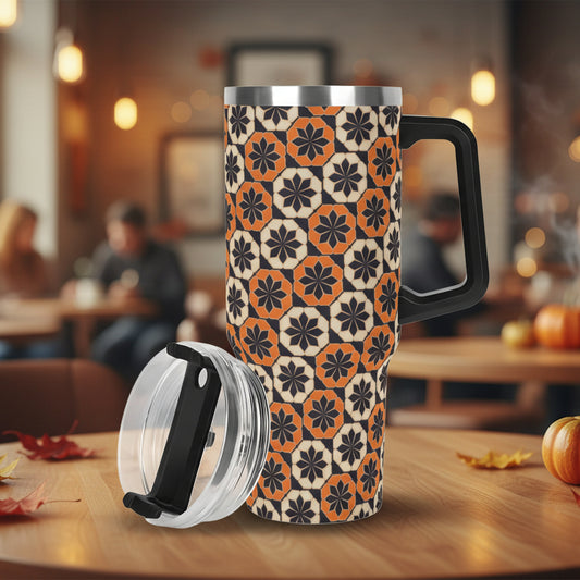 A product photo of an insulated stainless steel tumbler with a large black handle. The body of the cup is covered in a fall-themed repeating geometric design made of octagonal tile shapes in black, burnt orange, and cream. The tumbler's clear spill-proof lid with a black sliding closure rests detached on the white background next to it.