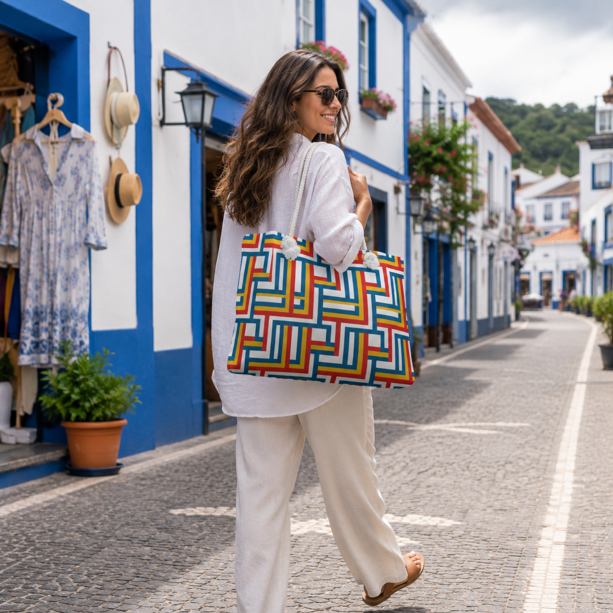 A woman walking down a cobblestone street carrying a large tote bag with a colorful geometric maze-like pattern in red, orange, yellow, green, blue, and purple.