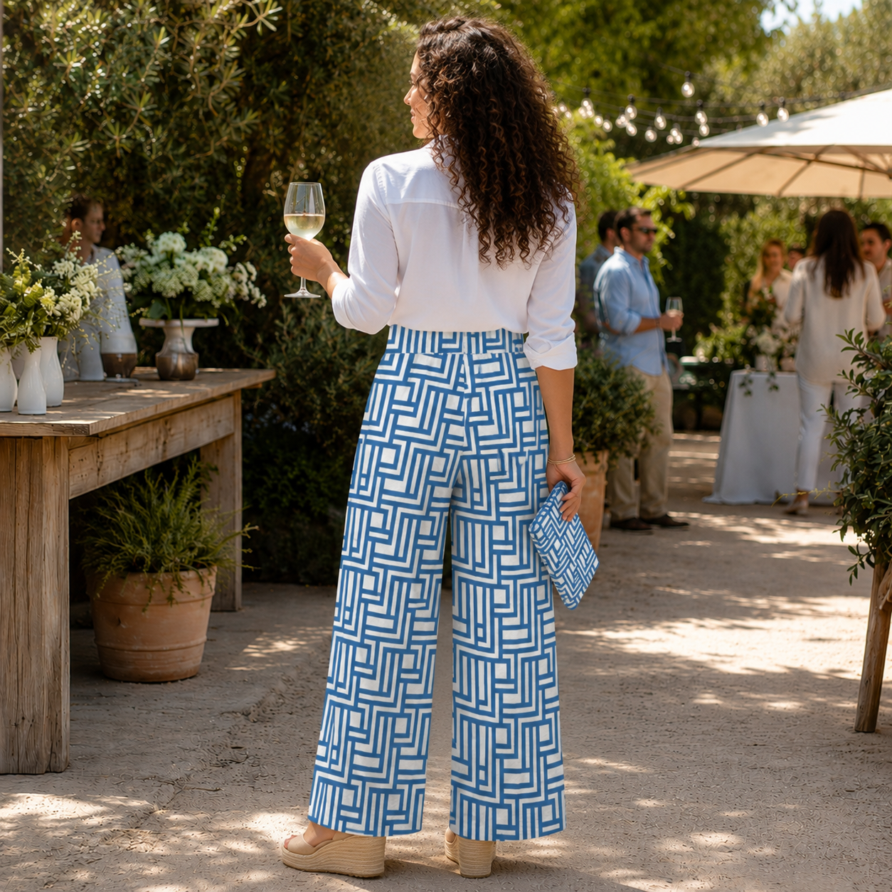 A woman stands outdoors at a garden party or event, shown from the back. She is wearing a white blouse and high-waisted wide-leg trousers with a bold blue and white geometric pattern, holding a wine glass and a matching blue clutch.