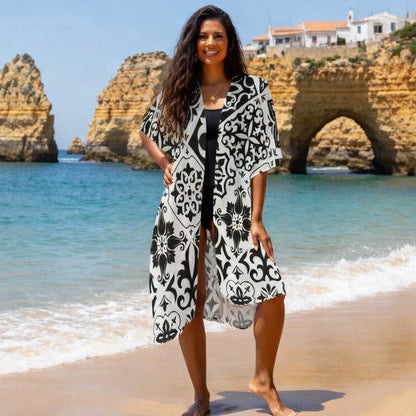 Woman in a black and white floral dress standing on a beach with rock formations and a house in the background.
