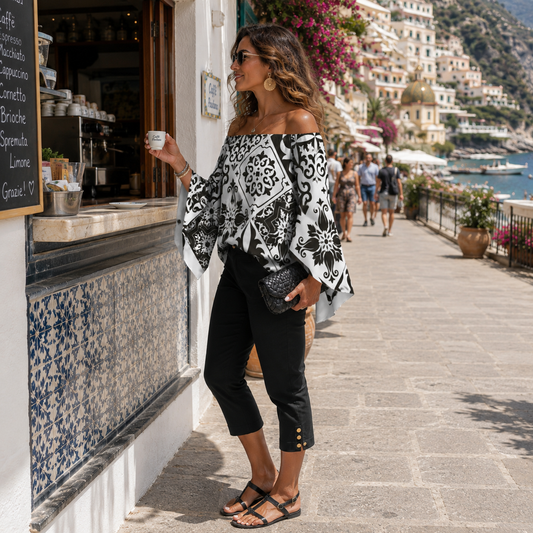 A woman stands on a sunny seaside promenade wearing an off-the-shoulder black and white patterned blouse, black cropped pants with gold buttons, and black sandals. She holds a small white cup near a cafe window, with a colorful coastal town and water in the background.