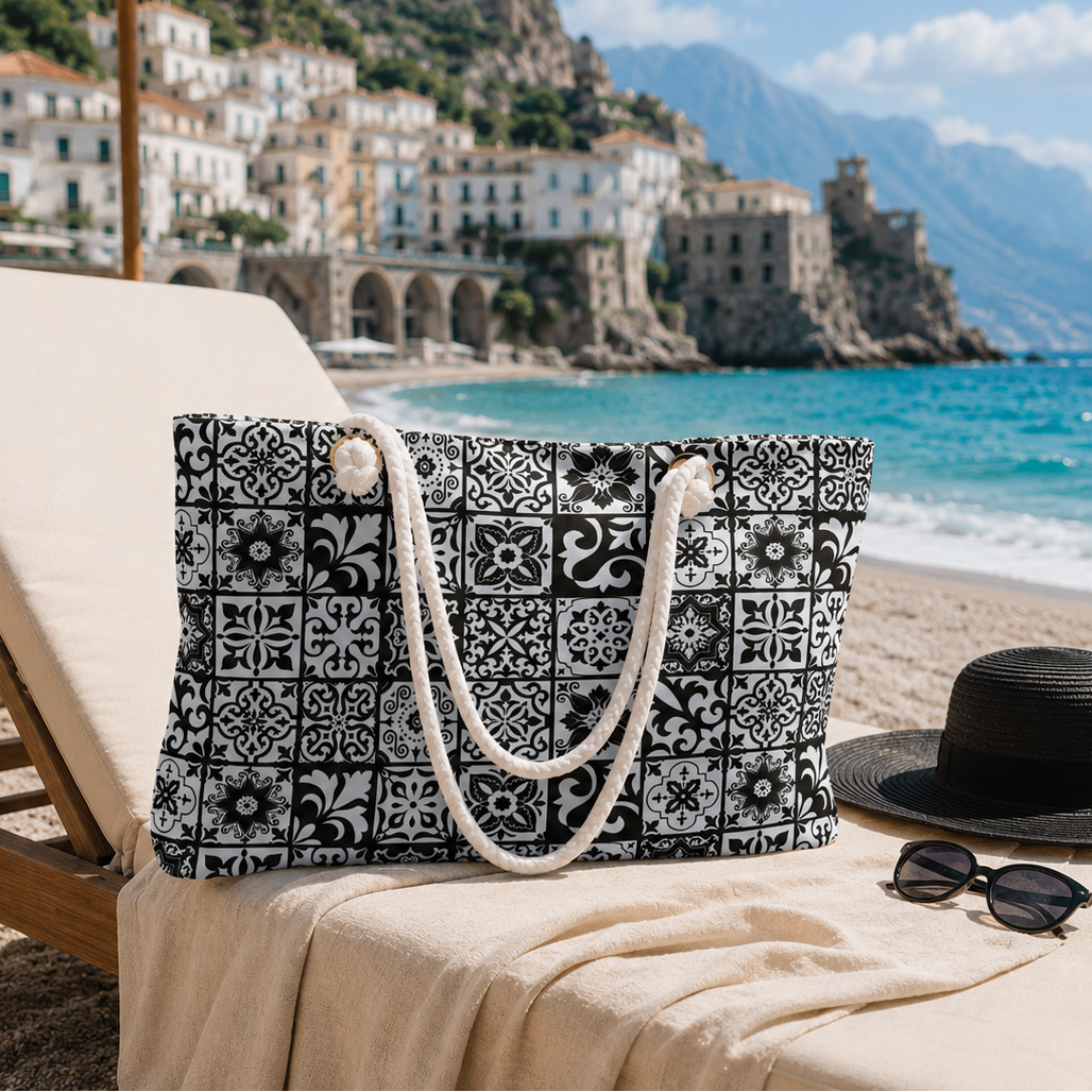 A black and white patterned tote bag with cream rope handles sits on a beige beach lounge chair at the beach, with a black sun hat, sunglasses, turquoise ocean, and coastal town in the background.