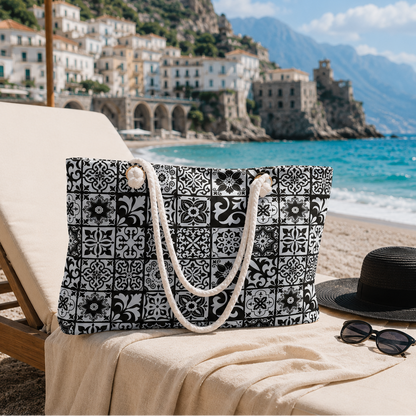 A black and white patterned tote bag with cream rope handles sits on a beige beach lounge chair at the beach, with a black sun hat, sunglasses, turquoise ocean, and coastal town in the background.