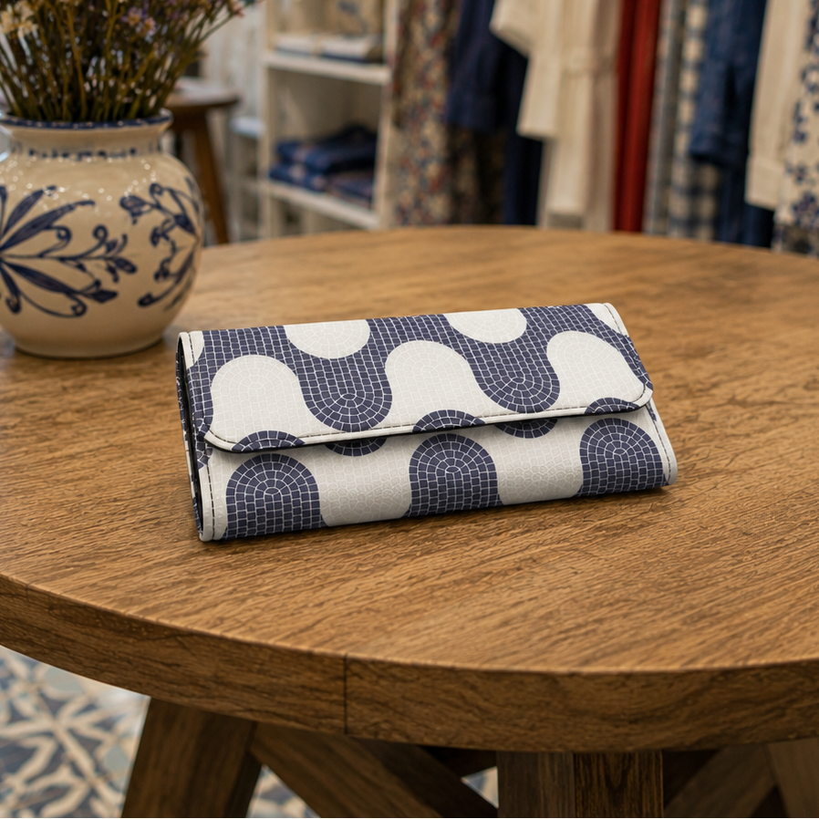A folded fabric clutch or wallet with a navy and white wavy geometric pattern sits on a round wooden table inside a boutique. A ceramic vase with dried flowers is on the left, and clothing racks are blurred in the background.