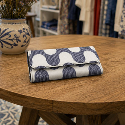 A folded fabric clutch or wallet with a navy and white wavy geometric pattern sits on a round wooden table inside a boutique. A ceramic vase with dried flowers is on the left, and clothing racks are blurred in the background.