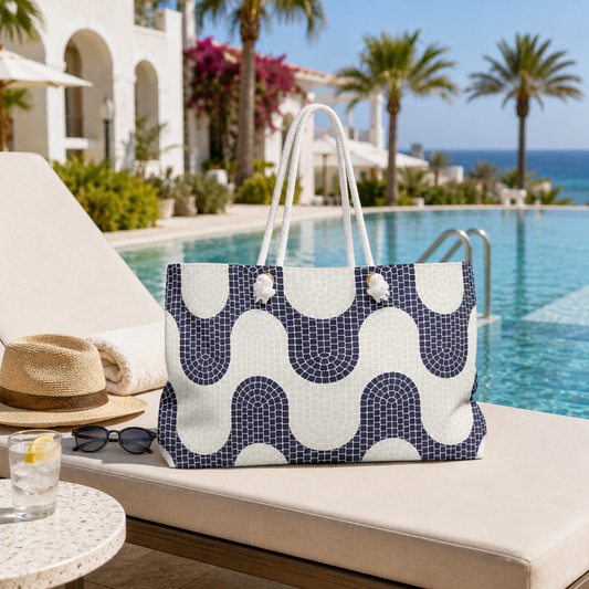 A navy and off-white geometric patterned tote bag with white rope handles sits on a beige sun lounger by a resort pool, with palm trees, a white building, and the ocean in the background.