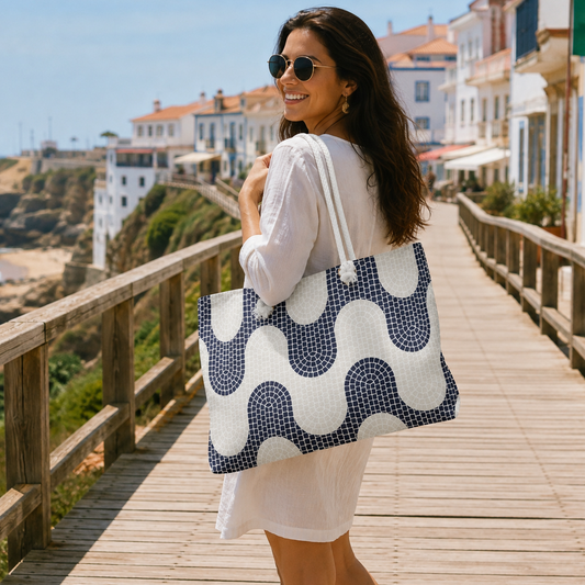 A woman wearing a white dress and sunglasses carries a large quilted tote bag with a navy and white wavy pattern while walking on a wooden boardwalk in a coastal town.