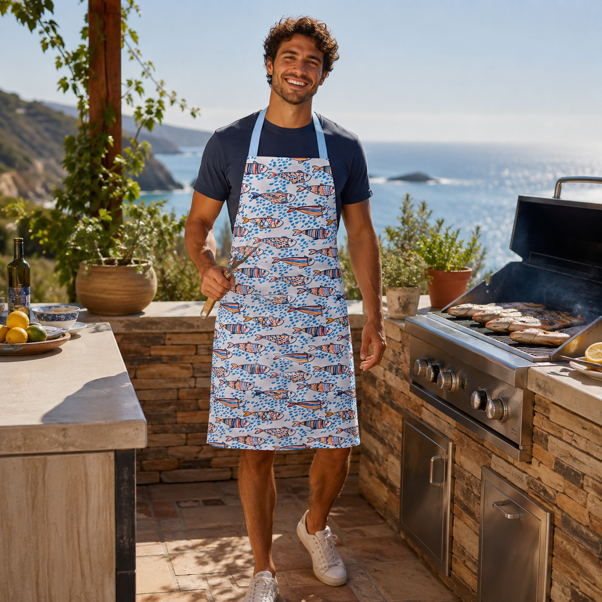 A man wearing a blue and white patterned apron stands on an outdoor patio next to a grill, holding tongs. The background features a scenic ocean view and a stone kitchen setup.