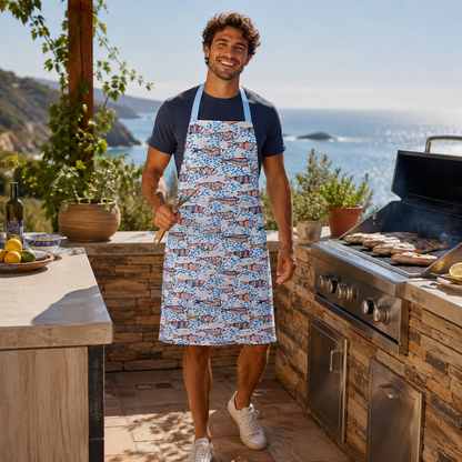 A man wearing a blue and white patterned apron stands on an outdoor patio next to a grill, holding tongs. The background features a scenic ocean view and a stone kitchen setup.