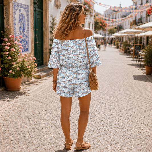 A woman wearing a light blue and white patterned off-the-shoulder romper walks down a sunny, cobblestone street lined with flowers and outdoor seating.