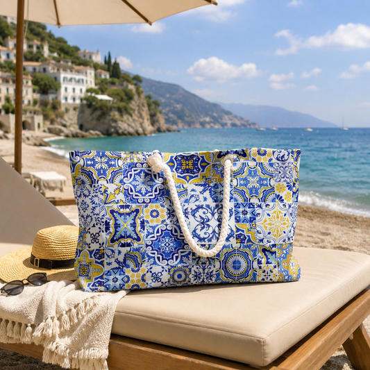 A large tote bag with a blue, white, and yellow Mediterranean tile pattern sits on a beige beach lounge chair at the seaside. A straw hat, sunglasses, and cream throw blanket are nearby, with the ocean, hills, and buildings in the background.