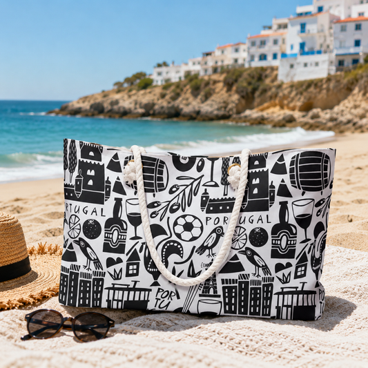 A black and white patterned tote bag with rope handles sits on a sandy beach next to a straw hat and sunglasses, with the ocean and coastal buildings in the background.