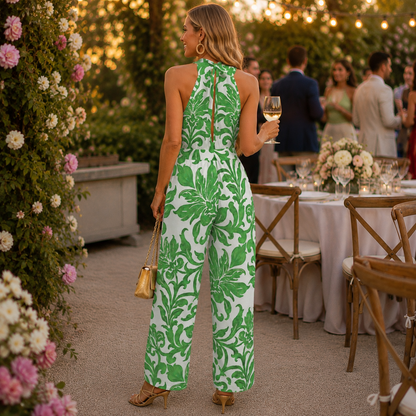 A woman stands with her back to the camera at an outdoor evening event, wearing a sleeveless white jumpsuit with a bold green floral pattern. She is holding a glass of white wine and carrying a small gold handbag.
