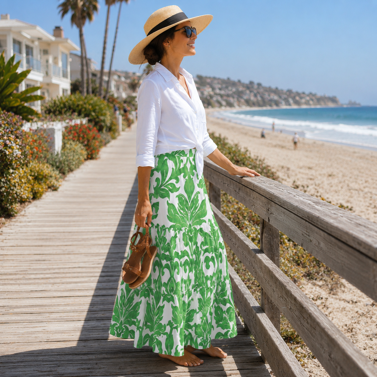 A woman stands on a wooden boardwalk by the beach wearing a white long-sleeve blouse and a green and white botanical print maxi skirt, with a straw hat and sunglasses.