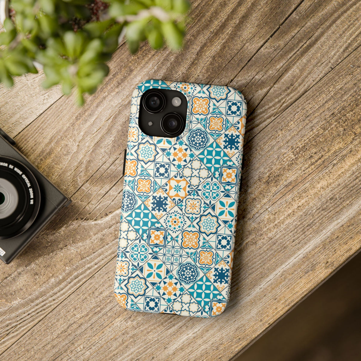 A phone case with a geometric mosaic pattern of turquoise, orange, and white Portuguese tiles, placed on a wooden surface next to a camera.