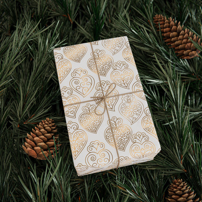 Gift box with gold heart pattern wrapped in paper, tied with string, on green foliage with pine cones.