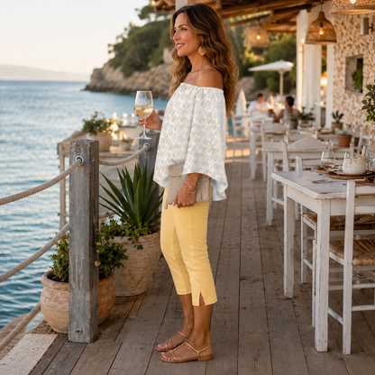 A woman stands on a seaside restaurant deck wearing a white off-the-shoulder top with a subtle geometric pattern and yellow cropped pants, holding a glass of white wine.