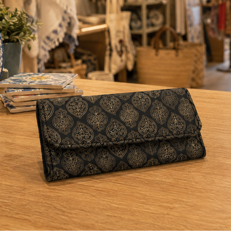 A dark patterned long wallet or clutch with a repeating ornate damask-style design, resting on a light wood table inside a cozy, softly blurred boutique.