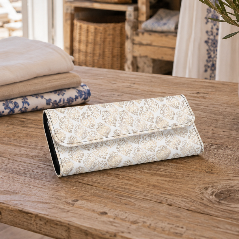 A white and beige patterned fold-over wallet or clutch rests on a rustic wooden table in a shop interior, with folded textiles, woven bags, and a beach scene visible through an open doorway in the background.