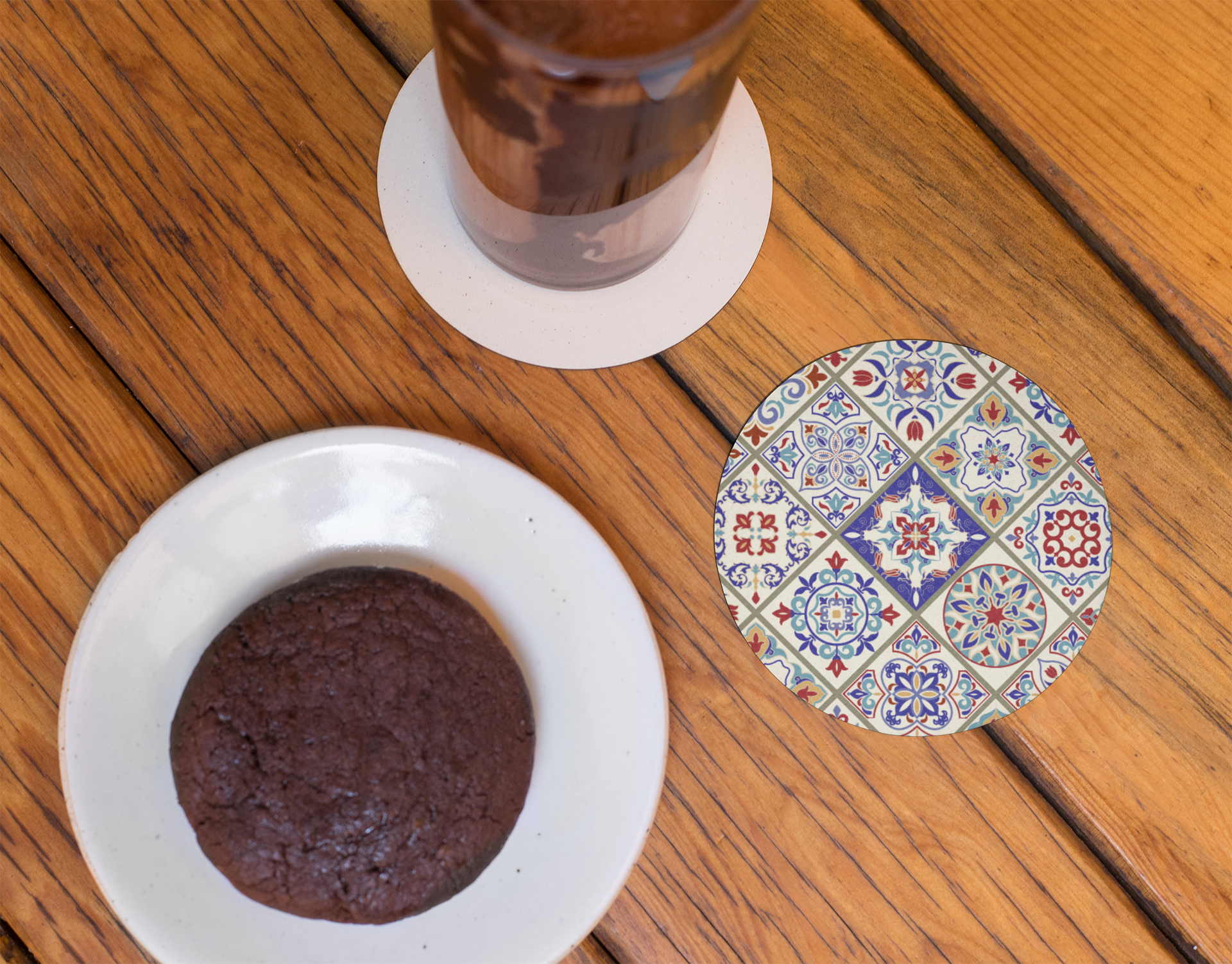 Chocolate cookie on a white plate with a colorful patterned coaster on a wooden surface.