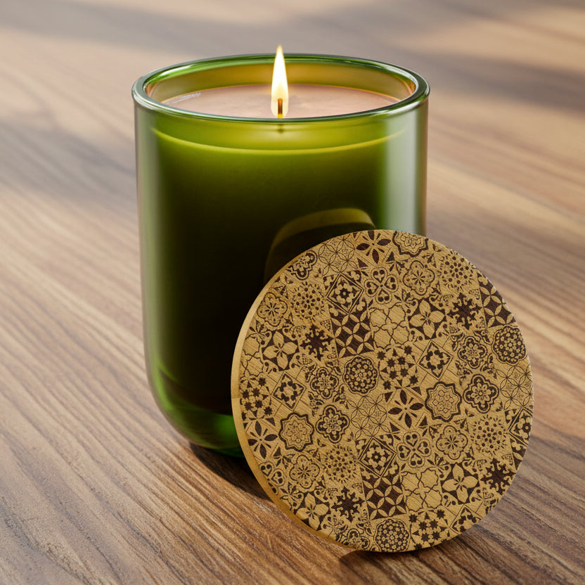 A lit green glass candle jar with a single flame, shown beside a round wooden lid featuring an intricate dark brown geometric and floral pattern, on a wooden table background.