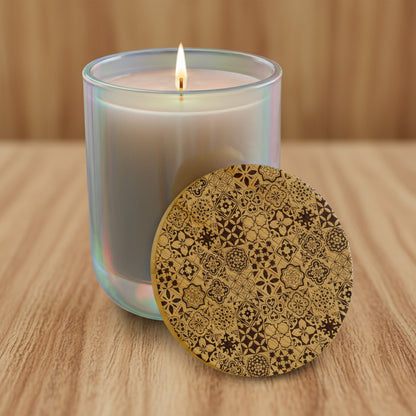A lit candle in a translucent iridescent glass jar with a small flame, shown beside a round wooden lid decorated with an intricate dark brown geometric floral pattern on a light wood background.