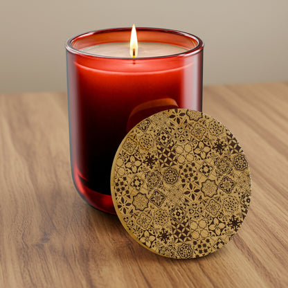 A lit red glass candle jar with a single flame, shown beside a round wooden lid or coaster featuring an intricate dark brown geometric and floral pattern, on a clean white background.