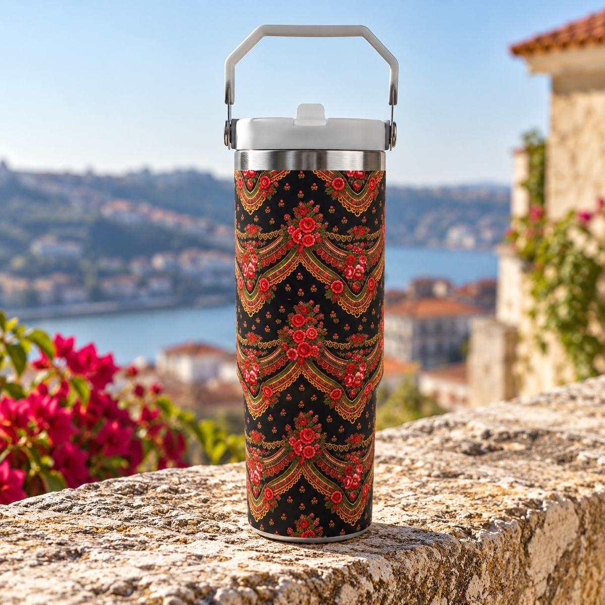 A tall insulated tumbler with a white handle and lid sits on a stone ledge outdoors. The tumbler body has a black background with an ornate red, gold, and orange decorative pattern.