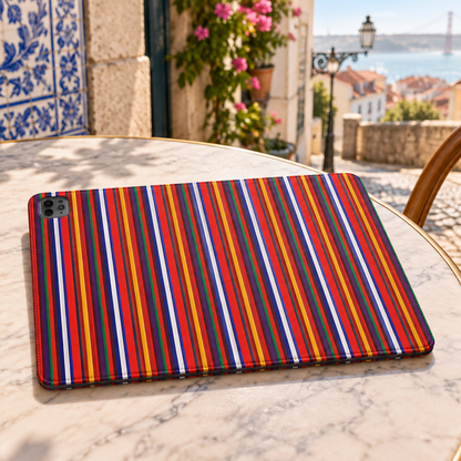 A tablet case with a vibrant vertical striped pattern in red, orange, yellow, blue, and white sits on a marble patio table. The setting is a sunny outdoor cafe with a cup of coffee, a blue and white ceramic vase of flowers, and a scenic view of a coastal city in the background.